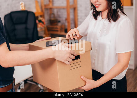 Asian lady leaving electronic signature on digital tablet and talking parcel from delivery man while working in office Stock Photo