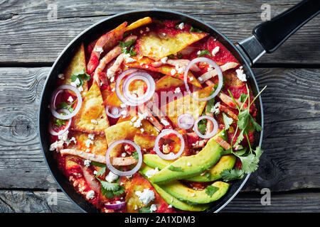 chilaquiles with ham, crumbled panela cheese, avocado slices and tomato salsa in a skillet on a rustic wooden table, mexican cuisine, horizontal view Stock Photo