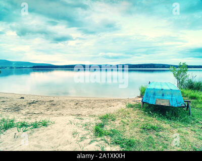 Blue fishing boat anchored on beach sand of lake. Smooth evening water level and calm. Large vallely of Lipno lake. Stock Photo