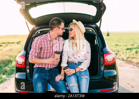 Cheerful young couple looking at mobile phone together while both sitting at the trunk of retro minivan with sea in the background Stock Photo