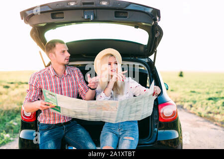 Beutiful people are sitting in trunk and talking to each other. Guy is holding a map and looking at girl while she is pointing on map and smiling. She Stock Photo