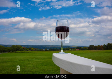 A glass of red wine photographed at on the arm rest of a white, plastic Adirondack chair near Cayuga Lake in the Finger Lakes Region of New York State Stock Photo