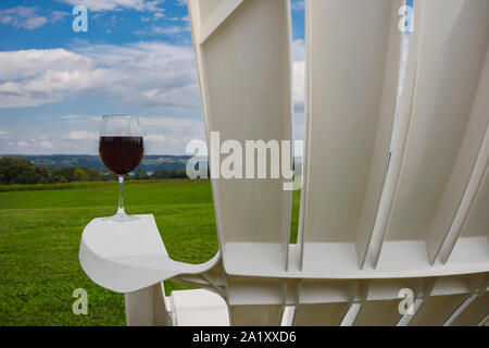 A glass of red wine photographed at on the arm rest of a white, plastic Adirondack chair near Cayuga Lake in the Finger Lakes Region of New York State Stock Photo