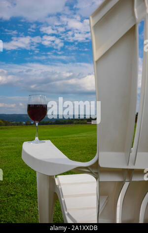 A glass of red wine photographed at on the arm rest of a white, plastic Adirondack chair near Cayuga Lake in the Finger Lakes Region of New York State Stock Photo