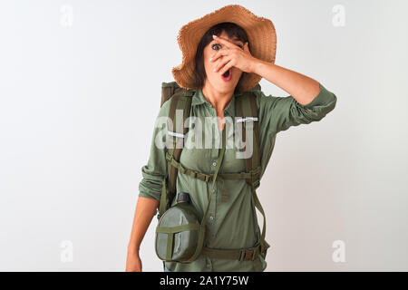 Hiker woman wearing backpack hat and water canteen over isolated white background peeking in shock covering face and eyes with hand, looking through f Stock Photo