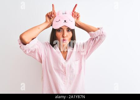 Young beautiful woman wearing sleep mask and pajama over isolated white background doing funny gesture with finger over head as bull horns