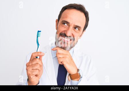 Middle age dentist man holding toothbrush standing over isolated white background serious face thinking about question, very confused idea Stock Photo
