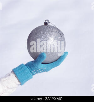 Woman's hand with blue knit glove holding big, silver Christmas ornament bauble with white snow background. Women's cold weather fashion accessories. Stock Photo