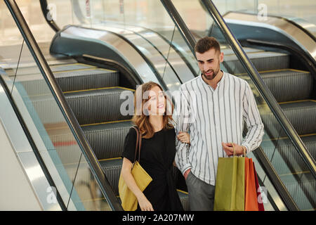 Smiling young couple with shopping bags standing on escalator after successful shopping in mall Stock Photo