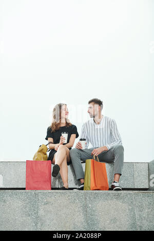 Positive young man and woman sitting on granite steps, drinking coffee and discussing sales and shopping Stock Photo