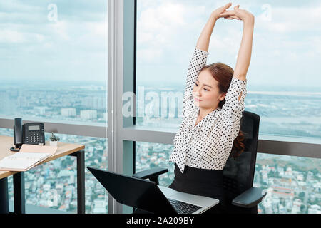 Beautiful young Asian businesswoman stretching arms after working on laptop in office all day long Stock Photo