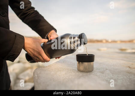 Man pours hot tea from a thermos into a cup which stands on an ice floe on the river bank in the spring sunny day Stock Photo