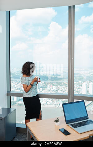 Pensive young Black businesswoman drinking cup of coffee and looking at big city through panoramic office window Stock Photo