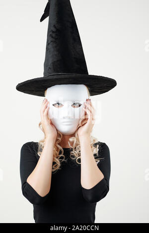 Portrait of young woman in witch costume putting on plastic white mask for Halloween party and looking at camera Stock Photo