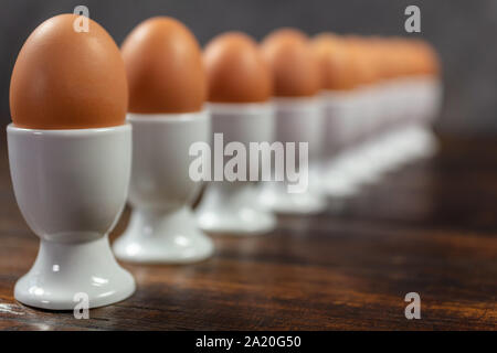 Ten boiled eggs in white egg cups in a line on a wooden table Stock Photo