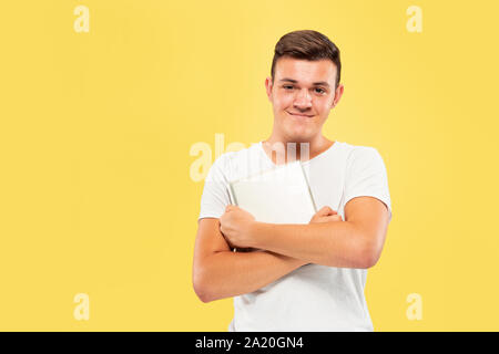 Caucasian young man's half-length portrait on yellow studio background. Beautiful male model in shirt. Concept of human emotions, facial expression, sales, ad. Holding tablet with hands crossed. Stock Photo