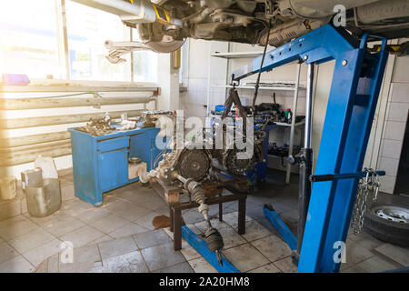 A car raised on a rotary lift for repair and under it a detached engine suspended on a blue crane near workbench in auto service. Stock Photo