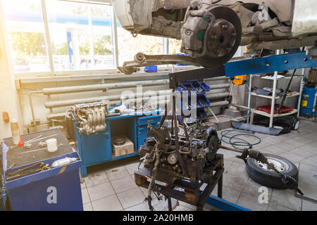 An old used car without wheel, raised on a lift for repair and under it a detached engine suspended on a blue crane near workbench in a vehicle repair Stock Photo