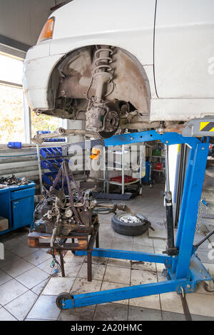 A white car raised on a lift for repair and under it a detached engine suspended on a blue crane on a lifting table in a vehicle repair industrial wor Stock Photo