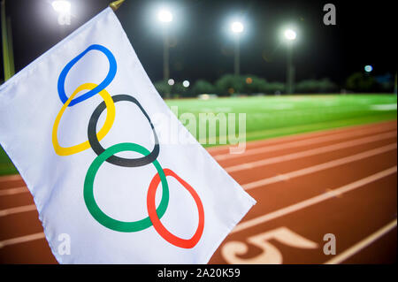 MIAMI, USA - AUGUST 15, 2019: An Olympic flag waves under the stadium floodlights of a red athletics track. Stock Photo