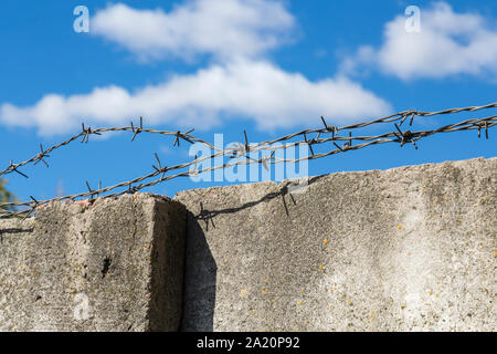 Three strands of barbed wire close-up, lying loosely on a fence of old concrete slabs with cracks and fungus against a blue sky and white clouds Stock Photo
