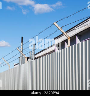 Barbed wire stretched in three threads over a metal fence of gray profiled sheet, industrial structure and chimneys behind the fence Stock Photo