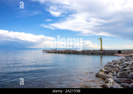 View of Issyk-Kul Lake, a mountain range with snow-capped peaks and a pier from a rocky beach. Kyrgyzstan Stock Photo