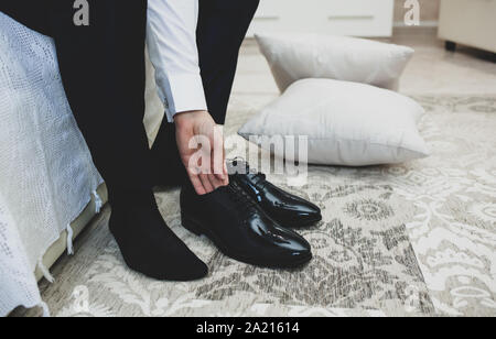 A man in a suit ties up shoelaces on black classic elegant shoes. Groom morning in hotel room before wedding ceremony. Stock Photo