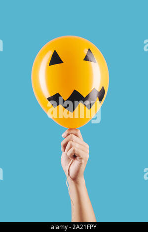 Scary face reminding jack-o-lantern on orange balloon in hand of person, isolated on light blue Stock Photo