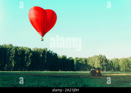 Young woman and man sitting on grass and looking at red hot air balloon in shape of heart. Love and future together concept. Stock Photo