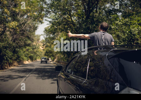 Young happy man drives a car and holds his hand out from the window. Driver enjoys driving and shows Ok sign with his hand out of window. Road trip, t Stock Photo