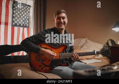 Portrait of a cheerful young man sitting on bed in front of window with american flag looking at camera Stock Photo