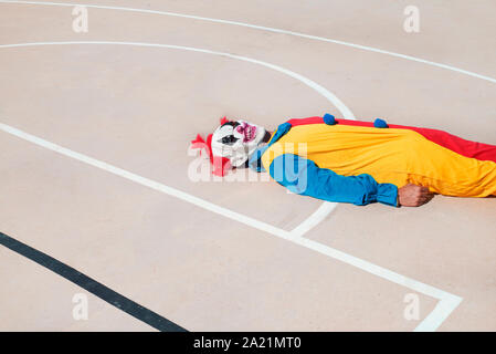 a scary clown, wearing a colorful yellow, red and blue costume, lying face up on an outdoor basketball court Stock Photo