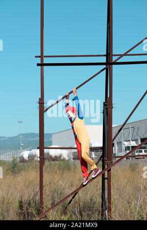 a scary clown wearing a colorful yellow, red and blue costume outdoors, hanging from the rusty structure of an abandoned billboard Stock Photo