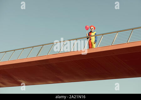 a creepy clown wearing a colorful yellow, red and blue costume, holding a red balloon in his hand, standing in a bridge outdoors Stock Photo