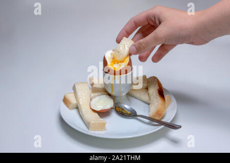 soft boiled chicken's egg, in an egg cup with buttered bread, soldiers, and a tea spoon resting on a plate. Hand dipping bread soldier into egg Stock Photo