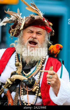 Pirate Day in Hastings. Close up head and shoulder of senior man dressed as Long John Silver with brushy grey beard, parrot on shoulder, eye-contact. Stock Photo