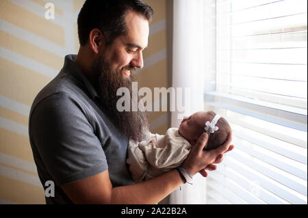Father smiling while holding newborn daughter at home by window