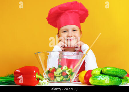 Beautiful little girl chef cook on a yellow background. A child is preparing a vegetable salad. Stock Photo