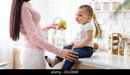 Tooth strengthening. Mom giving fresh apple to little baby son, sitting on kitchentop, panorama Stock Photo