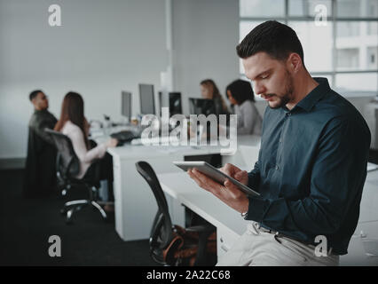 Portrait of young businessman leaning on desk using tablet pc and his colleagues working in the background Stock Photo