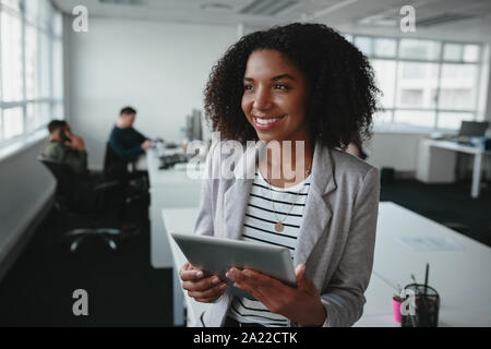 Portrait of a friendly successful young black businesswoman holding digital tablet in hand looking away at office Stock Photo