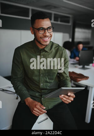 Thoughtful smiling young businessman holding digital tablet looking away while sitting at his working place in office Stock Photo