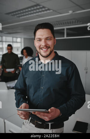 Portrait of a successful smiling young businessman holding digital tablet in hand looking at camera while coworkers having conversation on background Stock Photo