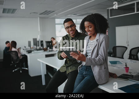 Successful two african american young businesspeople sitting on desk using digital tablet while colleague in background at office  Stock Photo