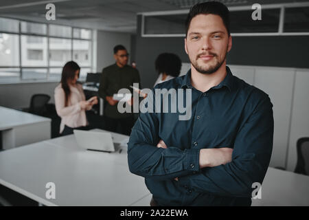 Handsome young professional man in office with folded arms looking at camera and other workers having conversation in background Stock Photo