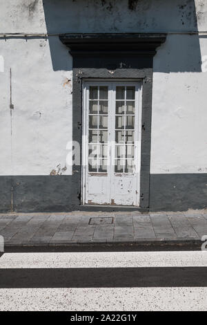 White door with a glass windows and a grill. Grey frame. Shadow of a balcony above. Part of a road with zebra crossing. Ribeira Grande, Sao Miguel, Az Stock Photo