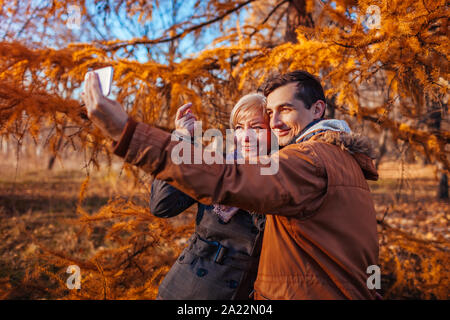 Adult son taking selfie with his mother using smartphone in autumn park. Family time Stock Photo