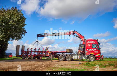 A red Herbies Haulage Ltd  Volvo FH13.520 crane lorry truck with low loader unloading farm machinery with its support legs and boom extended Stock Photo