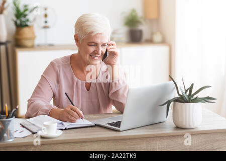Senior business woman working from home, discussing project with client, making notes in front of laptop, panorama with empty space Stock Photo
